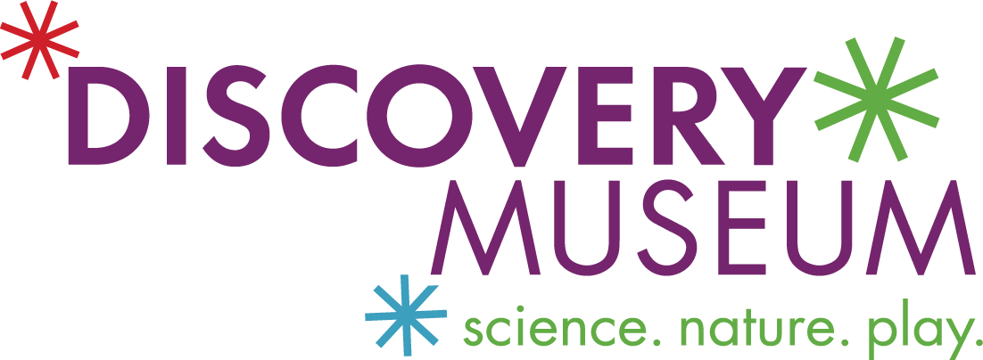 The image is a logo for the "Discovery Museum". The word "DISCOVERY" is in large, bold purple letters on top, and directly below in the same font but smaller size is the word "MUSEUM". The tagline "science. nature. play." is placed underneath the museum name in smaller green font, indicating the museum's focus areas.  The logo also features decorative elements in the form of stylized asterisks in red, blue, and green, positioned around the text, suggesting a sense of fun, wonder, and exploration that aligns with the museum's themes of science, nature, and play. The color scheme and design convey a friendly and educational environment suitable for all ages.