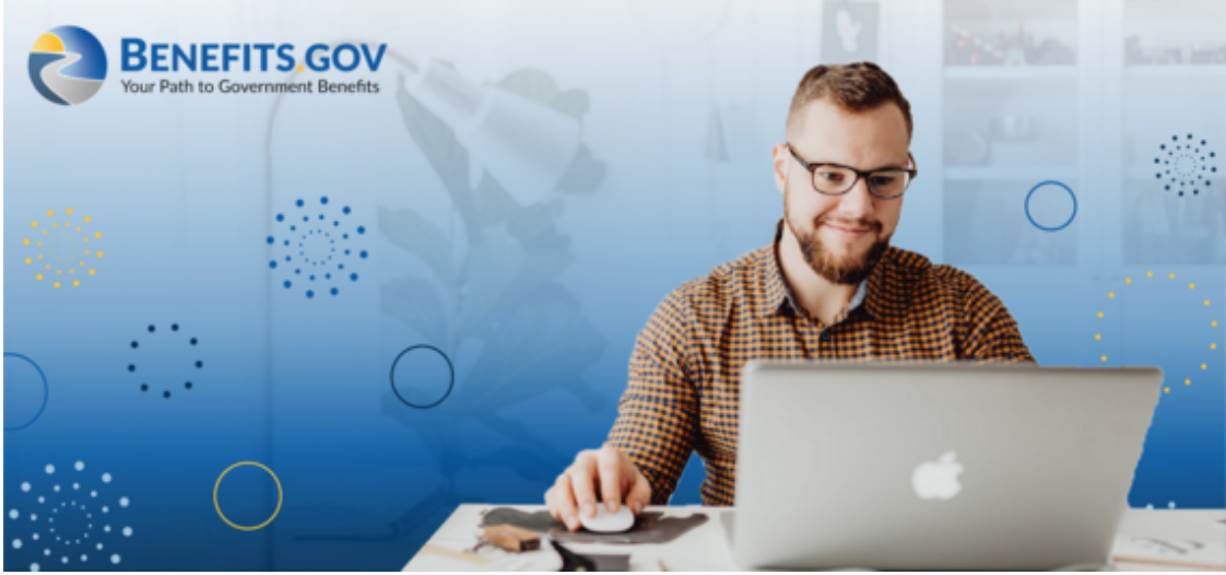 A white male with glasses sits with his laptop smiling on his cluttered desk. Behind him is a blue backdrop with gray faded images and multicolored circle patterns. At the top left is the benefits.gov logo.