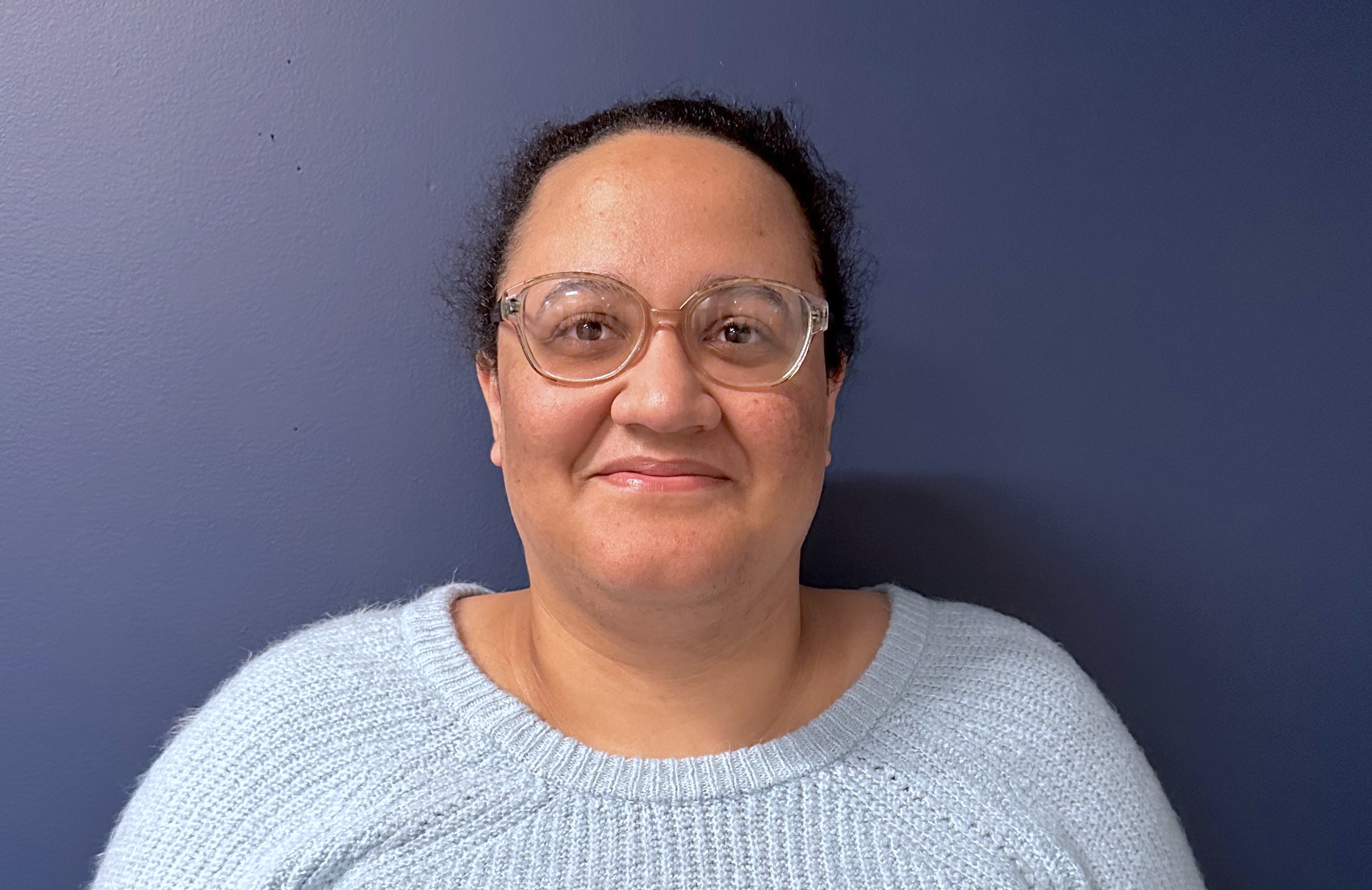 Image of a woman, with glasses and black hair tied back, she is wearing a white-ish blue sweater.
