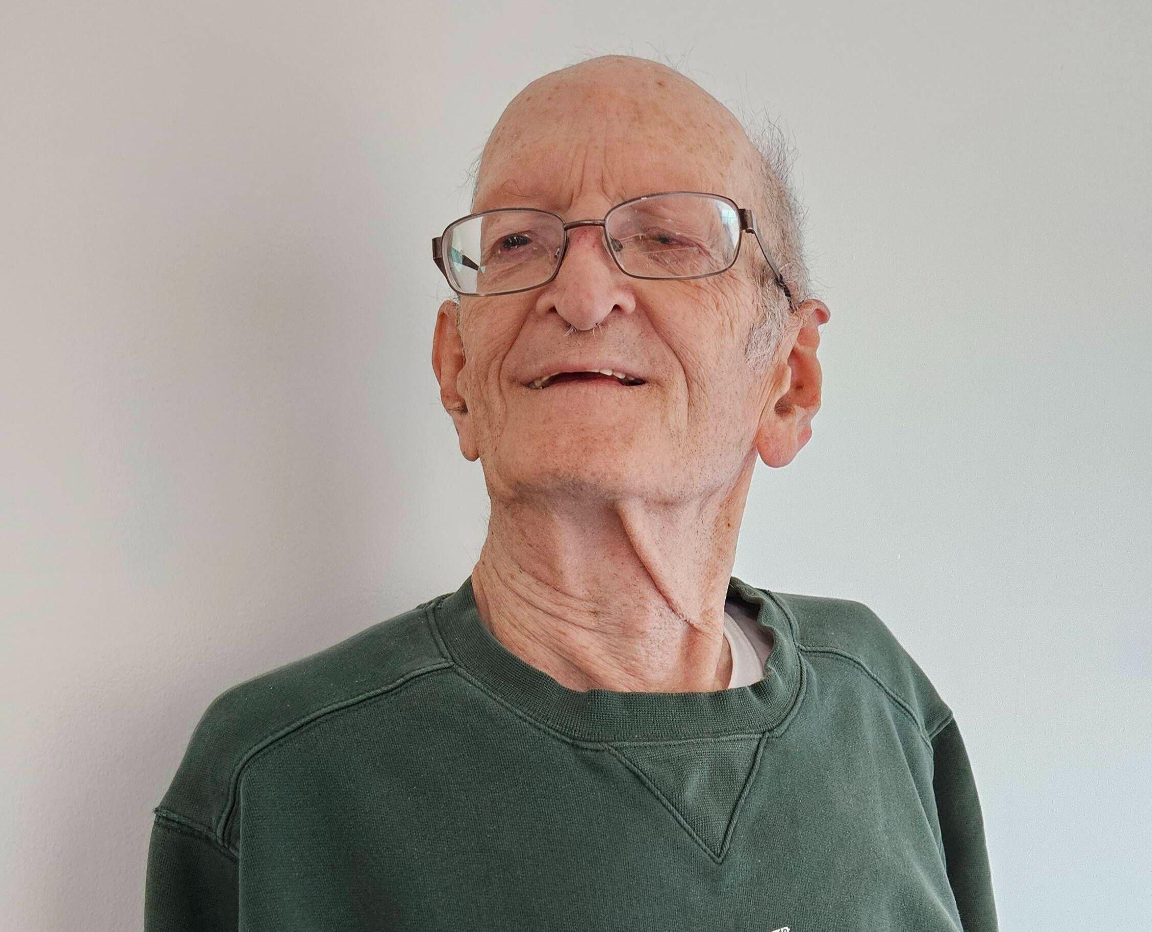 Image description: An image of Dennis Phillipe, a smiling elderly white man wearing a green sweater, and glasses.