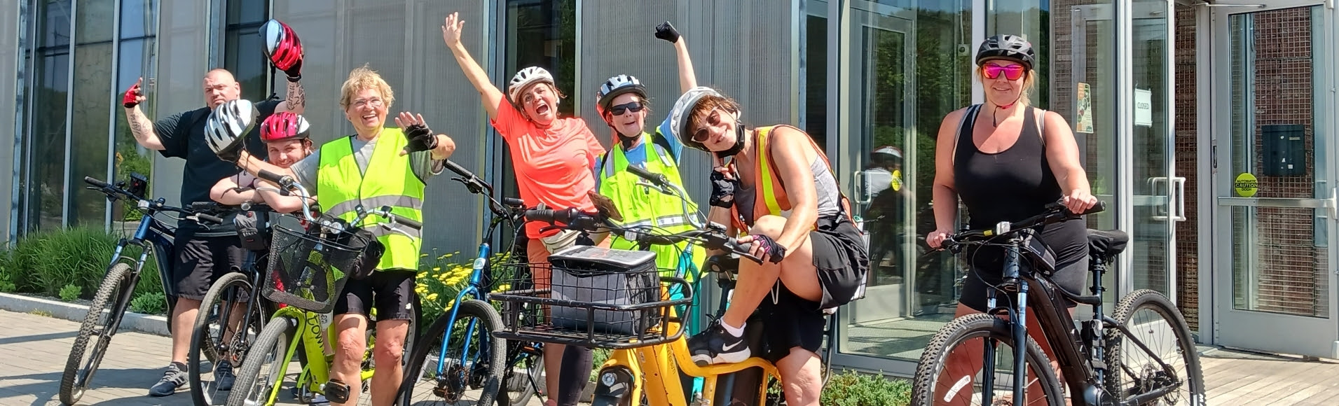 Image description:  7 people are lined up in front of a building, standing next to their bikes.  Some are wearing bike helmets, and some are holding their bike helmets in their hand. Some are dressed in tank tops, t-shirts and shorts and others are wearing bright reflective vests, sunglasses, and gloves smiling and waving at the camera. Below the picture is an application for the E-bike voucher.