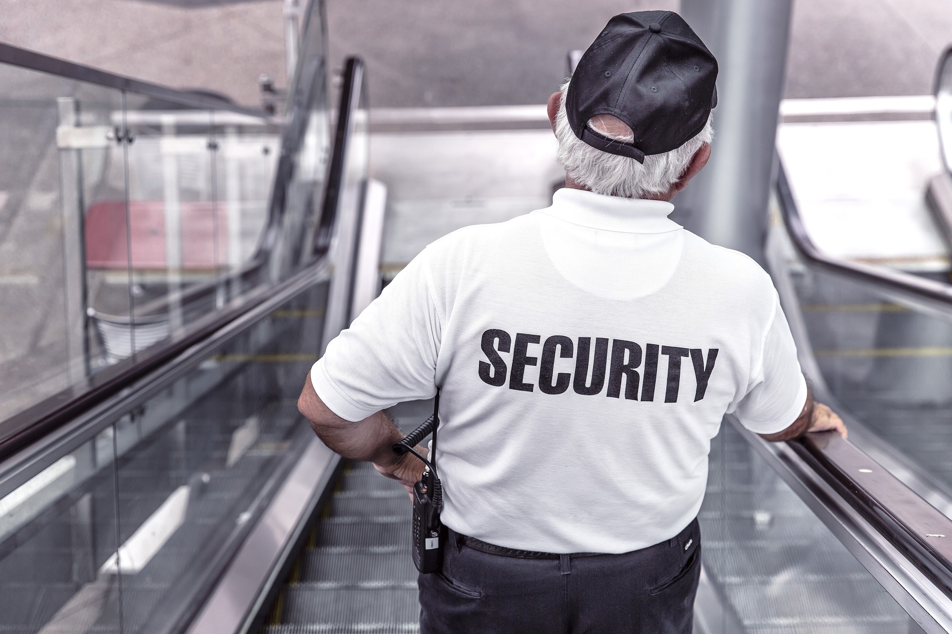 Image Description: A man in a white shirt with the word "security" printed in the back. declining down an escalader