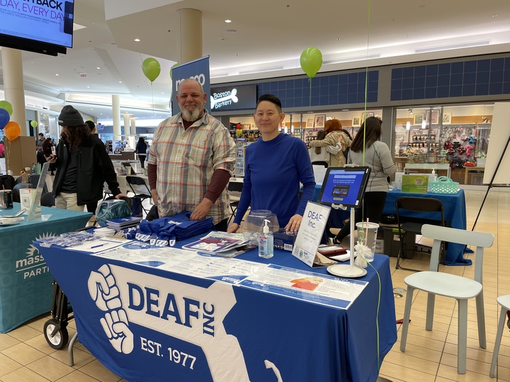 Image Description: Todd and Nayo at Peabody’s Health & Wellness Expo at Northshore Mall on March 30th, the two are standing at the table with the DEAF, Inc. logo draped over it. The table also has flyers, T-shirts, and items introducing others to DEAF, Inc. You can see people also in the background of them advertising other organizations. Image Description: Todd and Nayo at Peabody’s Health & Wellness Expo at Northshore Mall on March 30th, the two are standing at the table with the DEAF, Inc. logo draped over it. The table also has flyers, T-shirts, and items introducing others to DEAF, Inc. You can see people also in the background of them advertising other organizations.