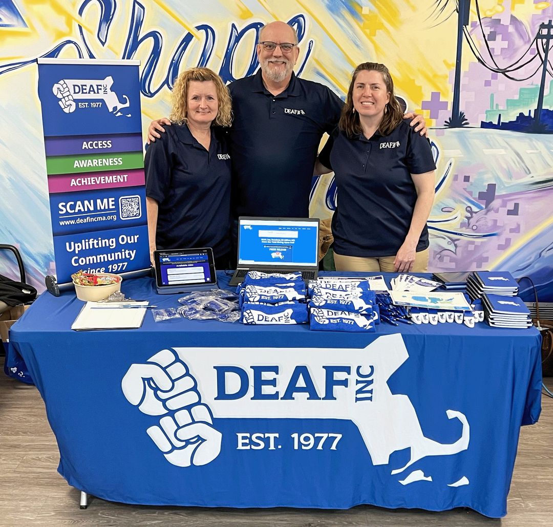 An image of a DEAF, Inc. table booth draped with a blue cloth showing off the organization’s logo and displaying an assortment of its’ merchandise. With DEAF, Inc’s staff Linda, Ronald, and Marie posing behind the table for the event in dark blue DEAF, Inc. shirts. An image of a DEAF, Inc. table booth draped with a blue cloth showing off the organization’s logo and displaying an assortment of its’ merchandise. With DEAF, Inc’s staff Linda, Ronald, and Marie posing behind the table for the event in dark blue DEAF, Inc. shirts.
