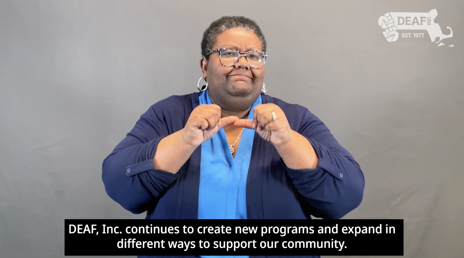 DEAF, Inc. Executive Director Darian Gambrell stands in front of a grey background wearing a light blue shirt and dark blue cardigan