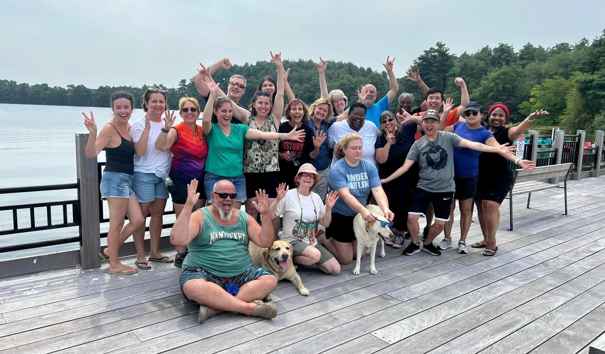 A group of smiling DEAF, Inc. staff, advocates, and service dogs at the Staff Appreciation day hosted by the DEAF, Inc. Board. They are all outside on a wooden dock by the lake with trees surrounding it.