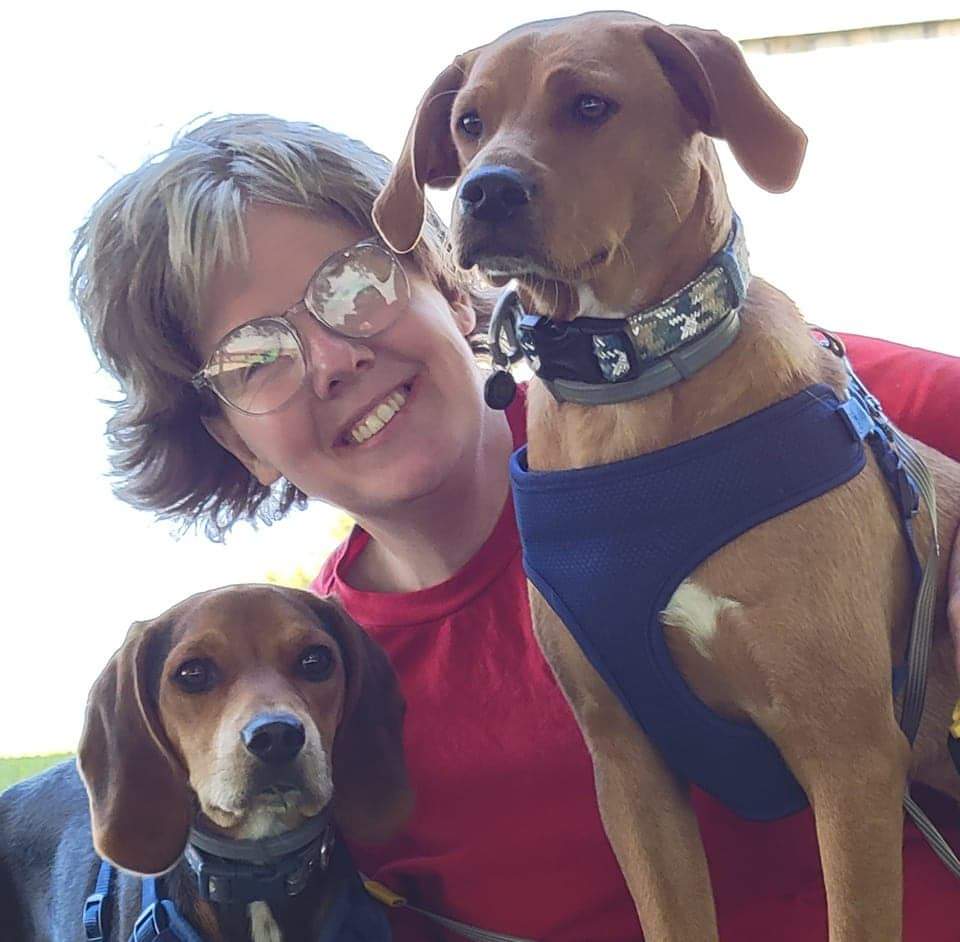 Photo of Samantha, a DEAF, Inc. Advocate, she is wearing a red shirt, glasses, and is smiling with her two brown dogs by her side.