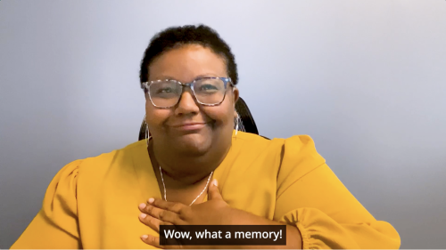 Thumbnail of Darian, DEAF, Inc.’s Executive Director, wearing a yellow blouse, white necklace, and glasses. She is signing with her hand to her chest. Caption at the bottom “Wow, what a memory!”