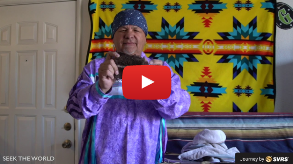 A YouTube still of a man in a purple sweater and bandanna holding up something of interest in front of a colorful cloth pattern.