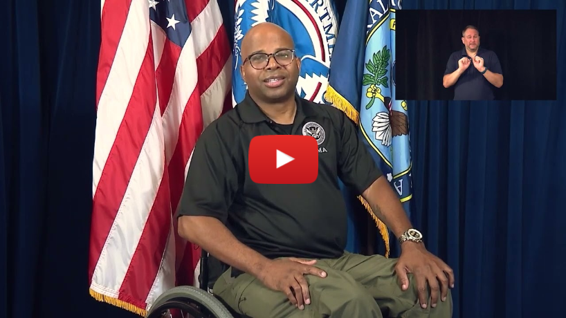Personal Preparedness: I Use a Wheelchair - A still frame from a video, featuring a man seated in a wheelchair in front of a backdrop of flags, including the American flag and other governmental flags. The man is wearing a black polo shirt with an official emblem, indicating he might be a representative of a government agency. Key Elements: The Man: He is bald, wearing glasses, and has a friendly expression, suggesting he is speaking or presenting information. Backdrop: The backdrop includes multiple flags, such as the American flag and possibly flags from the Department of Homeland Security or other related agencies. ASL Interpreter: There is an inset in the upper right corner showing an American Sign Language (ASL) interpreter signing, making the video accessible to the Deaf and Hard of Hearing community. Play Button: A large red play button with a YouTube logo is overlaid on the image