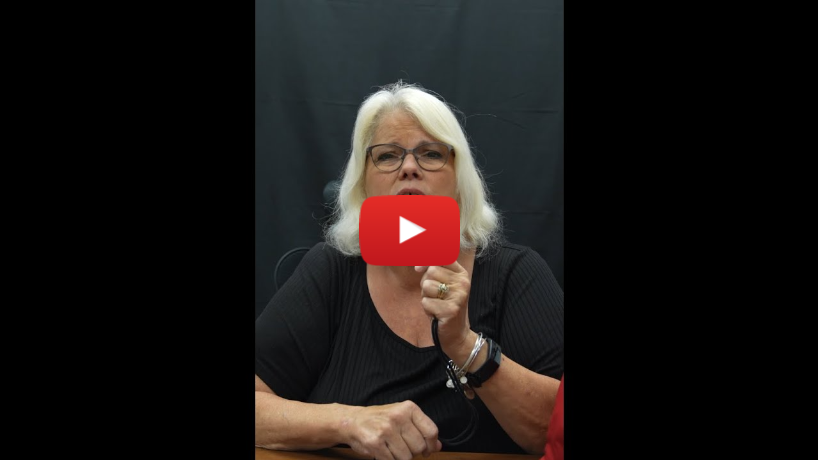Emergency Preparedness for the DeafBlind Community - A still frame from a video featuring a woman who appears to be delivering a message in American Sign Language (ASL). Key Elements: The Woman: She has shoulder-length white hair, is wearing glasses, and a black shirt. She is seated at a table and using ASL to communicate. Background: The background is solid black, which focuses attention on the woman. Play Button: A large red play button with the YouTube logo is overlaid on the image, indicating that this is a screenshot from a video from YouTube.