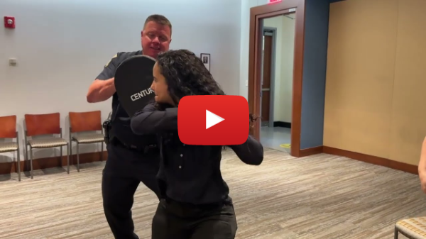 Visual description:  Rossy is a Hard of Hearing brown skinned Hispanic female with black eyes, thick dark brown curly hair.  She is wearing a dark blouse. She is in a room where a uniformed policeman is holding a strike pad.  She is practicing hitting the strike pad with her elbow twice then turning around, holding both arms up indicating that she is strong.